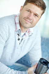 Image showing Portrait of man smiling and sitting on couch holding a flask. Attractive male inside his home with a travel coffee mug in his hand. Handsome caucasian male drinking tea and relaxing in an apartment