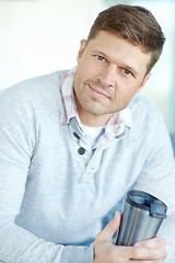 Image showing Portrait of man smiling and sitting on couch holding a flask. Attractive male inside his home with a travel coffee mug in his hand. Handsome caucasian male drinking tea and relaxing in an apartment