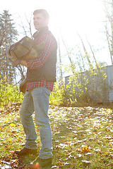 Image showing One male lumberjack holding wooden logs after chopping tree in remote landscape. Serious focused adult man in autumn wear standing alone outside, carrying firewood from the forest