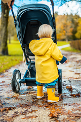 Image showing Sun always shines after the rain. Small blond infant boy wearing yellow rubber boots and yellow waterproof raincoat walking in puddles, pushing stroller in city park, holding mother's hand after rain.