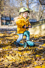 Image showing Adorable toddler boy wearing yellow protective helmet riding baby scooter outdoors on autumn day. Kid training balance on mini bike in city park. Fun autumn outdoor activity for small kids.