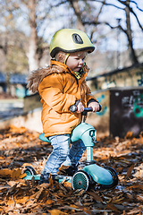 Image showing Adorable toddler boy wearing yellow protective helmet riding baby scooter outdoors on autumn day. Kid training balance on mini bike in city park. Fun autumn outdoor activity for small kids.