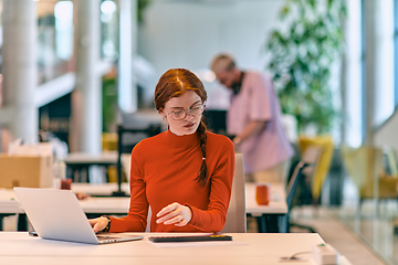 Image showing In a modern startup office, a professional businesswoman with orange hair sitting at her laptop, epitomizing innovation and productivity in her contemporary workspace.