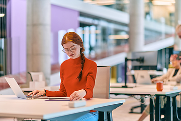 Image showing In a modern startup office, a professional businesswoman with orange hair sitting at her laptop, epitomizing innovation and productivity in her contemporary workspace.