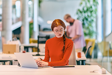 Image showing In a modern startup office, a professional businesswoman with orange hair sitting at her laptop, epitomizing innovation and productivity in her contemporary workspace.