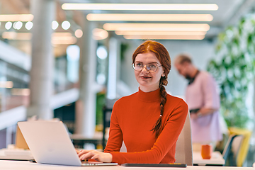 Image showing In a modern startup office, a professional businesswoman with orange hair sitting at her laptop, epitomizing innovation and productivity in her contemporary workspace.