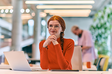 Image showing In a modern startup office, a professional businesswoman with orange hair sitting at her laptop, epitomizing innovation and productivity in her contemporary workspace.