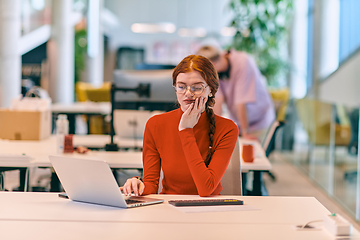 Image showing In a modern startup office, a professional businesswoman with orange hair sitting at her laptop, epitomizing innovation and productivity in her contemporary workspace.