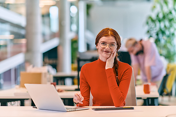 Image showing In a modern startup office, a professional businesswoman with orange hair sitting at her laptop, epitomizing innovation and productivity in her contemporary workspace.