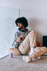 Image showing African-American entrepreneur taking a relaxing break from work, sitting on the floor while using wireless headphones and a smartphone for some digital entertainment.