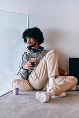 Image showing African-American entrepreneur taking a relaxing break from work, sitting on the floor while using wireless headphones and a smartphone for some digital entertainment.