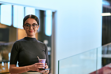 Image showing In a bustling modern office a businesswoman in glasses juggles her tasks, sipping coffee and using her smartphone, epitomizing the dynamic and multitasking nature of contemporary corporate life