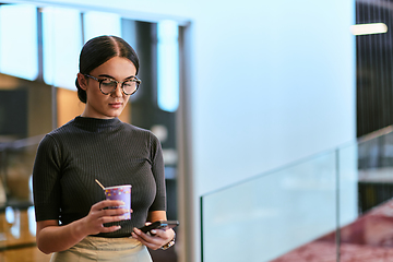 Image showing In a bustling modern office a businesswoman in glasses juggles her tasks, sipping coffee and using her smartphone, epitomizing the dynamic and multitasking nature of contemporary corporate life