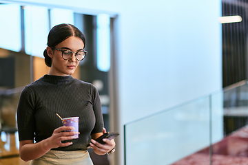 Image showing In a bustling modern office a businesswoman in glasses juggles her tasks, sipping coffee and using her smartphone, epitomizing the dynamic and multitasking nature of contemporary corporate life