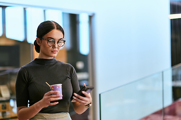 Image showing In a bustling modern office a businesswoman in glasses juggles her tasks, sipping coffee and using her smartphone, epitomizing the dynamic and multitasking nature of contemporary corporate life