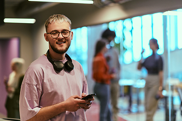 Image showing In a contemporary startup office, a modern blond entrepreneur stands, engrossed in his smartphone, epitomizing the dynamic and tech-savvy essence of the modern business world.