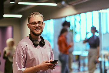 Image showing In a contemporary startup office, a modern blond entrepreneur stands, engrossed in his smartphone, epitomizing the dynamic and tech-savvy essence of the modern business world.