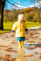 Image showing Sun always shines after the rain. Small bond infant boy wearing yellow rubber boots and yellow waterproof raincoat walking in puddles in city park on sunny rainy day.