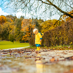 Image showing Sun always shines after the rain. Small bond infant boy wearing yellow rubber boots and yellow waterproof raincoat walking in puddles in city park on sunny rainy day.