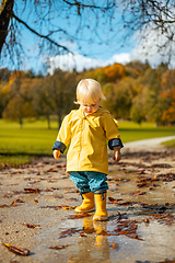 Image showing Sun always shines after the rain. Small bond infant boy wearing yellow rubber boots and yellow waterproof raincoat walking in puddles in city park on sunny rainy day.