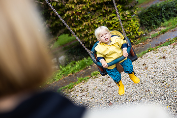 Image showing Mother pushing her infant baby boy child wearing yellow rain boots and cape on swing on playground outdoors on cold rainy overcast autumn day in Ljubljana, Slovenia