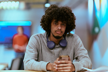 Image showing In a modern startup office, an African American man, balancing work and technology, utilizes headphones and a smartphone, emblematic of contemporary multitasking and productivity