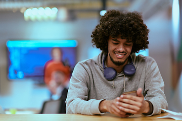 Image showing In a modern startup office, an African American man, balancing work and technology, utilizes headphones and a smartphone, emblematic of contemporary multitasking and productivity