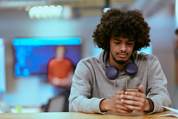 Image showing In a modern startup office, an African American man, balancing work and technology, utilizes headphones and a smartphone, emblematic of contemporary multitasking and productivity