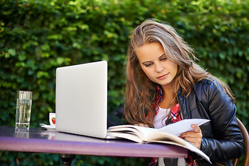 Image showing Woman, book and reading for research in cafe, outdoors and sitting for college, work or assignment. Young, female student and technology for online, submission or document by laptop with internet