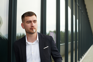 Image showing Amidst the corporate hustle, a modern businessman in a black suit takes a smoke break outside his workplace, seeking a moment of relaxation in the midst of a busy day.