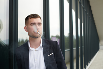 Image showing Amidst the corporate hustle, a modern businessman in a black suit takes a smoke break outside his workplace, seeking a moment of relaxation in the midst of a busy day.