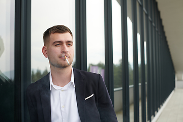 Image showing Amidst the corporate hustle, a modern businessman in a black suit takes a smoke break outside his workplace, seeking a moment of relaxation in the midst of a busy day.