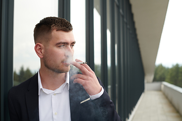 Image showing Amidst the corporate hustle, a modern businessman in a black suit takes a smoke break outside his workplace, seeking a moment of relaxation in the midst of a busy day.