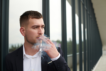 Image showing Amidst the corporate hustle, a modern businessman in a black suit takes a smoke break outside his workplace, seeking a moment of relaxation in the midst of a busy day.