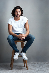 Image showing Fashion, serious and portrait of man in studio on a stool with casual, cool and stylish outfit. Confidence, handsome and young male model from Mexico with trendy style on chair by gray background.