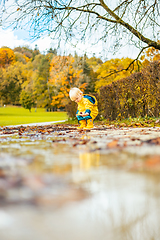 Image showing Sun always shines after the rain. Small bond infant boy wearing yellow rubber boots and yellow waterproof raincoat walking in puddles in city park on sunny rainy day.