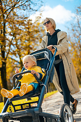 Image showing Young beautiful mother wearing a rain coat pushing stroller with her little baby boy child, walking in city park on a sunny autumn day