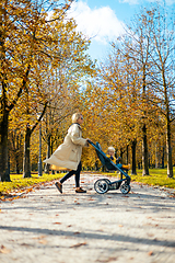 Image showing Young beautiful mother wearing a rain coat pushing stroller with her little baby boy child, walking in city park on a sunny autumn day