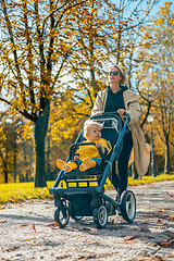 Image showing Young beautiful mother wearing a rain coat pushing stroller with her little baby boy child, walking in city park on a sunny autumn day