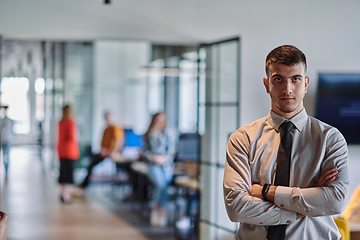 Image showing A young business leader stands with crossed arms in a modern office hallway, radiating confidence and a sense of purpose, embodying a dynamic and inspirational presence.