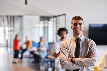 Image showing A young business leader stands with crossed arms in a modern office hallway, radiating confidence and a sense of purpose, embodying a dynamic and inspirational presence.