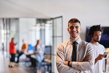 Image showing A young business leader stands with crossed arms in a modern office hallway, radiating confidence and a sense of purpose, embodying a dynamic and inspirational presence.