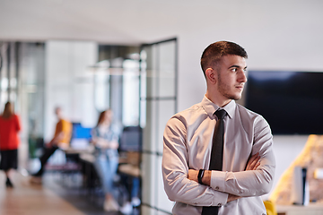 Image showing A young business leader stands with crossed arms in a modern office hallway, radiating confidence and a sense of purpose, embodying a dynamic and inspirational presence.