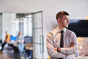 Image showing A young business leader stands with crossed arms in a modern office hallway, radiating confidence and a sense of purpose, embodying a dynamic and inspirational presence.