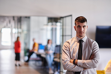 Image showing A young business leader stands with crossed arms in a modern office hallway, radiating confidence and a sense of purpose, embodying a dynamic and inspirational presence.