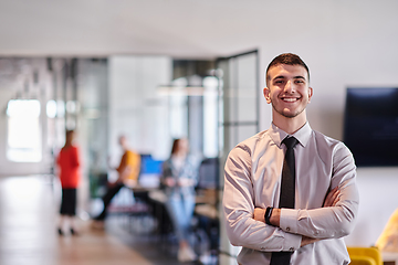 Image showing A young business leader stands with crossed arms in a modern office hallway, radiating confidence and a sense of purpose, embodying a dynamic and inspirational presence.