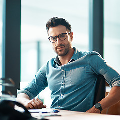 Image showing Young, confident and professional business man wearing glasses, sitting at his desk and working in a modern office portrait. A casual and serious corporate male in the finance department