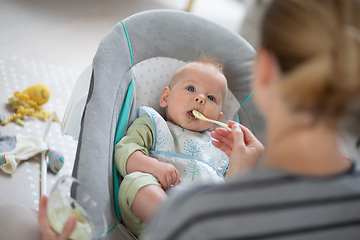 Image showing Mother spoon feeding her baby boy infant child in baby chair with fruit puree. Baby solid food introduction concept.