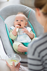 Image showing Mother spoon feeding her baby boy infant child in baby chair with fruit puree. Baby solid food introduction concept.