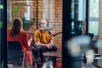 Image showing A gathering of young business professionals, some seated in a glass-walled office, engage in a lively conversation and record an online podcast, embodying modern collaboration and dynamic interaction